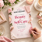 A pink planner titled 'The Ultimate Beauty & Self-Care Planner' by Secret Solutions is styled on a marble surface with flowers, a candle, a cup of coffee, makeup items, and a hand writing in the cover with a pink pen.