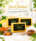 Three bars of Secret Solutions Lemon Turmeric & Kojic Acid Brightening Soap arranged on a marble slab with lemons, turmeric roots, and bowls of ingredients around them. The brand logo and name are displayed at the top of the image.