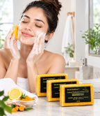 A woman with face wash foam on her face applies brightening soap from three bars of Secret Solutions Lemon Turmeric & Kojic Acid soap, shown in a bright bathroom setting with a lemon slice and turmeric root on the counter.