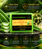 A green rectangular bar of Secret Solutions Natural Cool Fresh Aloe Soap Bar is centered on a dark stone surface, surrounded by aloe vera leaves and gel slices. The product has a black label with green and orange text.