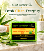 A rectangular bar of Secret Solutions Cool Fresh Aloe Soap Bar is displayed on a marble surface with sliced aloe vera leaves, aloe gel, and a white bottle in the background. The soap bar has a green textured wrapper with a black and orange label.